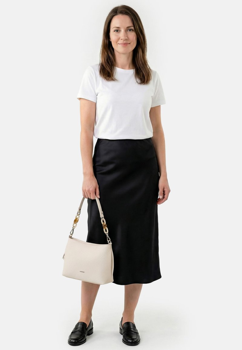 Nainen valkoisessa lyhythihaisessa paidassa ja mustassa midihameessa, pitäen käsissään beigeä käsilaukkua, jossa on kilpikonnakuviolliset ketjulisäkkeet, mustat loafersit jalassa ja yksivärinen tausta.