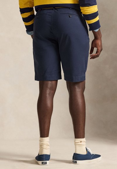 Man wearing navy blue tailored shorts, yellow and navy striped sweater, cream socks, and navy sneakers, standing against plain background.