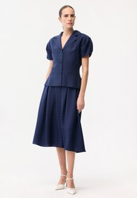 Une femme se tient debout, portant un blazer bleu marine à manches courtes boutonné et une jupe midi à larges jambes assortie, avec des escarpins blancs à bout pointu.