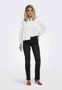 Hvit skjorte med knapper og plisserte detaljer på skuldrene, sammen med tettsittende svarte jeans og svarte høye sandaler, satt mot en lys bakgrunn.