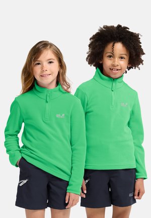 Due bambini indossano pile verdi brillanti con collo a zip, con un logo. Entrambi abbinano le magliette a pantaloncini blu scuro. Tessuto morbido e testurizzato.