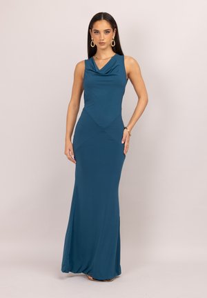 Une femme porte une robe longue jusqu’au sol, bleu canard, avec un décolleté drapé et des bijoux en or, sur un fond de studio uni.