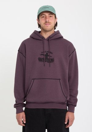 Jeune homme portant un sweat à capuche violet avec un motif d'épouvantail et une casquette vert clair, debout devant un fond blanc uni.