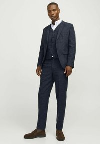 Marineblauer, maßgeschneiderter Dreiteiler mit strukturiertem Stoff, schmal geschnittenen Hosen und einem einreihigen Blazer. Braune Wildlederschuhe ergänzen das Outfit.