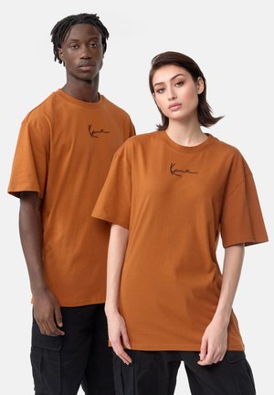 Zwei Modelle tragen übergroße, braune Baumwoll-T-Shirts mit schwarzen gestickten Logos. Die T-Shirts haben einen Rundhalsausschnitt und kurze Ärmel.