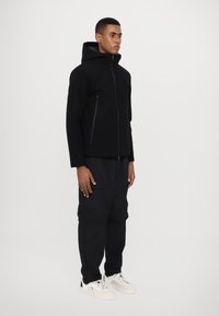 Svart jakke med glidelås og hette, med ensfarget tekstur og to sidelommer; kombinert med løstsittende svarte bukser og hvite joggesko.