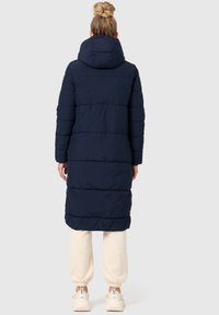 Navy puffer coat with a hood, featuring a quilted design and full-length sleeves. The hem has a diagonal pattern. Paired with light pants.