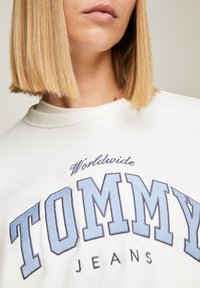 Detailní záběr na osobu, která má na sobě bílý top s jemně modrým vyšitým nápisem "TOMMY JEANS" a textem "Worldwide" pod bradou.