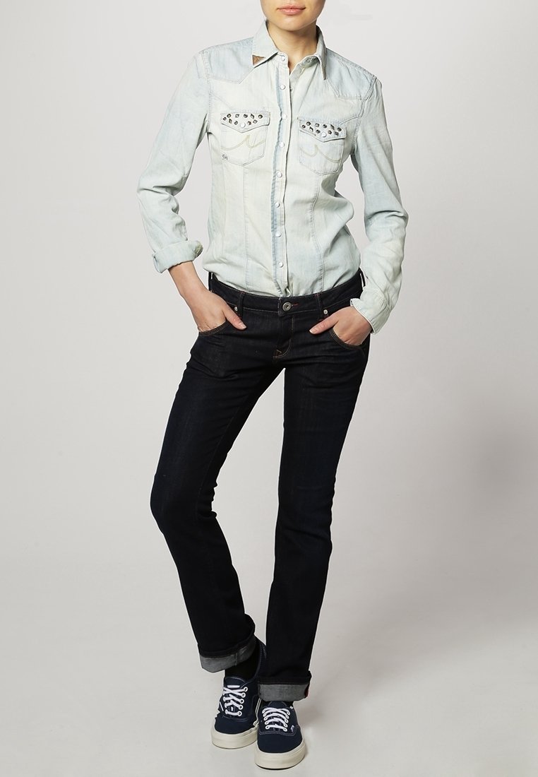 Camisa de mezclilla azul clara con tachuelas en los bolsillos, botonadura frontal y mangas largas. Combinada con jeans oscuros y zapatillas navy.