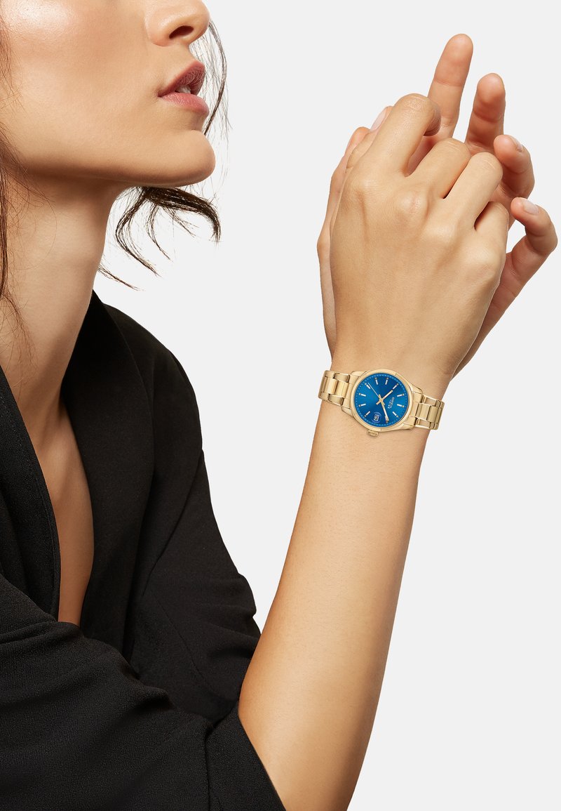 Montre-bracelet dorée avec un cadran bleu, bracelet en acier inoxydable, boîtier rond et fonction date. Main positionnée élégamment sur un fond en tissu noir.