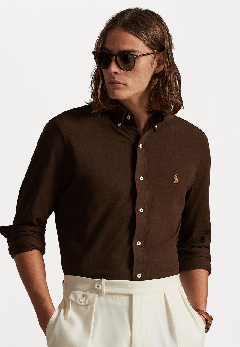 Καφέ πουκάμισο με κουμπιά και μακριά μανίκια, λευκά κουμπιά και μικρό χρυσό λογότυπο. Σε συνδυασμό με κρεμ πλισέ παντελόνι.