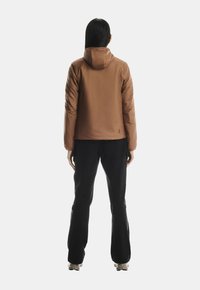 Persoană văzută din spate, purtând o jachetă cu glugă maro, pantaloni negri și încălțăminte bej, stând pe un fundal alb simplu.