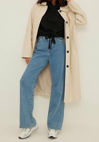 Cappotto lungo beige, felpa con cappuccio nera, jeans a gamba larga blu chiaro e sneakers bianche con dettagli neri. Tessuto morbido e vestibilità casual.