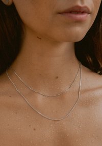 Schichtige silberne Ketten, eine glatt und die andere mit Struktur. Die Haut wirkt leicht feucht, mit sichtbaren Wassertropfen auf dem Schlüsselbein.
