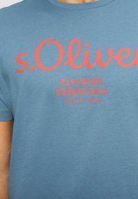Blaues Rundhals-T-Shirt aus weichem Baumwollstoff mit rotem Text: "s.Oliver", der prominent angezeigt wird, darunter "ELEVATED ESSENTIALS" und "SINCE 1969".