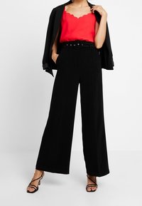 Rode tanktop met scalloped halslijn, zwarte hoge taillebroek met wijde pijpen en een zwarte blazer, gecombineerd met strappy zwarte hakken.