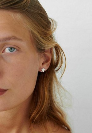 Naeripilt naisest, kellel on helepruunid juuksed ja kes kannab väikseid hõbedasi lillekujulisi kõrvarõngaid ühetaolise heleda tausta ees.