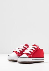 Zapatillas de lona rojas con cordones blancos y puntera. Suela a rayas en blanco y negro; parte inferior de goma texturizada. Diseño pequeño y bajo.