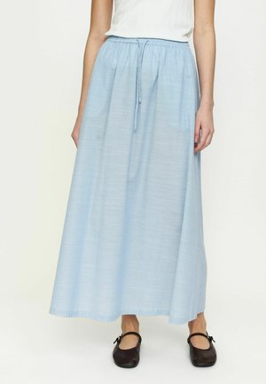 Kvinne iført en lys blå ankel-lang skjørt med snor i livet, hvit topp og svarte Mary Jane-sko, stående på en ensfarget bakgrunn.