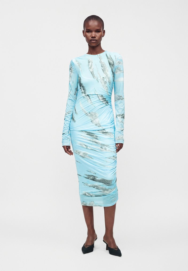 Femme portant une robe midi bleu clair à manches longues avec des motifs abstraits argentés et des chaussures noires à talons bas, debout sur fond blanc.