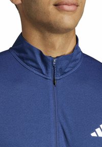 Blauwe sporttop met een hoge kraag en een korte rits. Heeft een gestructureerde stof en een wit logo aan de linkerkant.