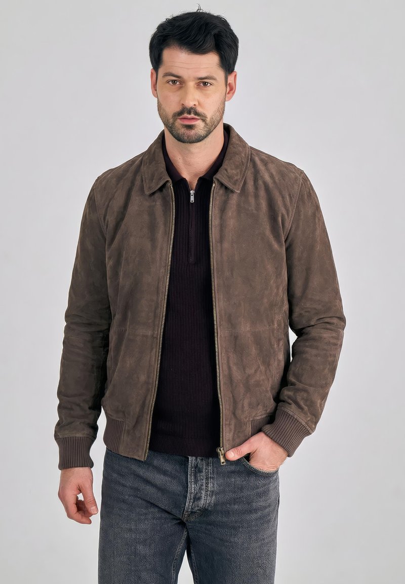 Mies, jolla on ruskea mokkatakki mustan vetoketjullisen neulepaidan ja sinisten farkkujen päällä, seisoo yhdellä kädellä taskussa yksinkertaisen taustan edessä.