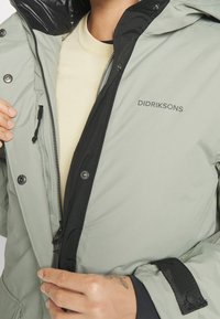 Veste isolante vert clair avec accents noirs, fermeture éclair à l'avant, boutons-pression, et le logo "DIDRIKSONS" sur la poitrine. Poignets ajustables.