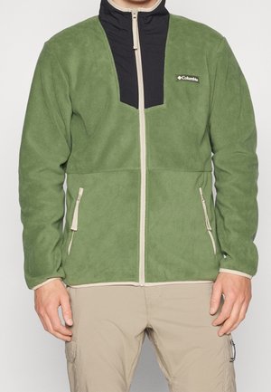 Mężczyzna w zielonej polarowej kurtce Columbia z beżowym zamkiem błyskawicznym i czarnym kołnierzem, w zestawie z beżowymi spodniami.