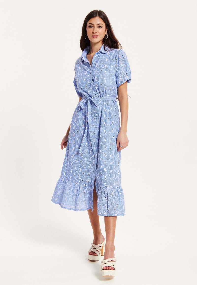 Light blue shirt dress with short sleeves, a cinched waist, ruffled hem, and white geometric pattern. Paired with chunky white sandals.