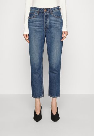 Mujer con vaqueros azul denim de pierna recta con botón y cremallera en el frente, combinados con tacones negros de punta afilada y un top blanco de manga larga.