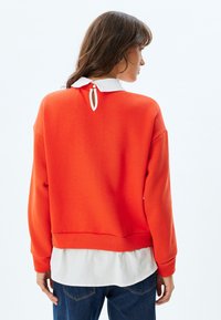 Sudadera naranja hecha de tela suave con un cuello blanco por debajo. Presenta un detalle de ojal en la parte posterior y puños acanalados.