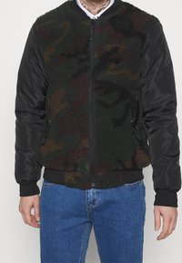 Man i en fleecejacka med kamouflage-mönster och svarta nylonärmar, stående mot en enfärgad bakgrund.