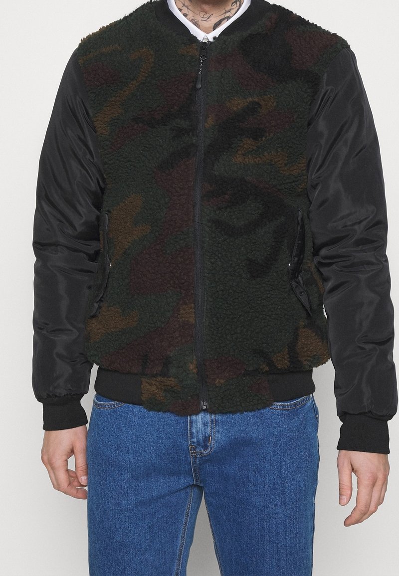 Man i en fleecejacka med kamouflage-mönster och svarta nylonärmar, stående mot en enfärgad bakgrund.