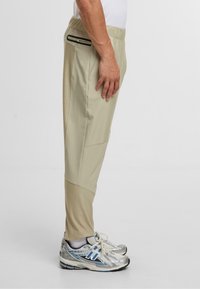 Beżowe spodnie sportowe z elastycznym pasem, boczną kieszenią na zamek, zwężanymi nogawkami i kontrastowymi panelami, w połączeniu z srebrnymi sneakersami.