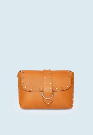 Bolso de mano de cuero marrón con tachuelas plateadas y una correa frontal con un anillo plateado, sobre un fondo azul claro.