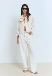 Beige denimová vesta se zapínáním na druky, bílá průsvitná halenka s volánkovými rukávy, široké bílé kalhoty a ploché sandály. Nosené tmavé sluneční brýle.