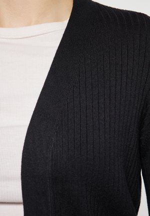 Černý žebrovaný cardigan s hlubokým výstřihem ve tvaru V, vyrobený z texturované látky. Následně přeložený přes bílý přiléhavý top s tenkými pruhy.