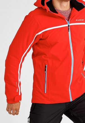 Homme portant une veste à capuche rouge vif avec des bandes blanches sur les manches et une fermeture éclair bleue, assortie à un pantalon noir.