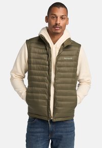Olivengroene gewatteerde vest met horizontale stiksels, voorrits en hoge kraag, gelaagd over een crèmekleurige hoodie, gecombineerd met blauwe jeans.