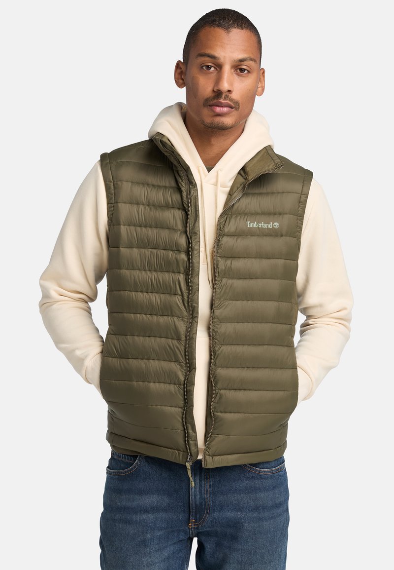 Olivengroene gewatteerde vest met horizontale stiksels, voorrits en hoge kraag, gelaagd over een crèmekleurige hoodie, gecombineerd met blauwe jeans.
