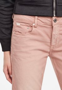 Detailní záběr na osobu v světle růžových džínách se stříbrným knoflíkem a zipem, viditelný rukáv černé bundy a levá ruka odpočívající na boku.