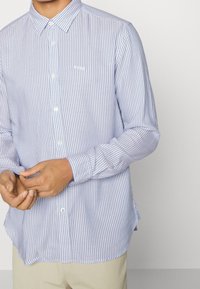 Ljusblå och vitrandig skjorta med knappar och långa ärmar, bröstlogo och rundad nederkant. Tillverkad av ett mjukt tyg.