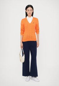 Camisola laranja de malha com decote em V, camisa branca com colarinho por baixo, combinada com calças largas azuis-escuras e ténis brancos. Segurando uma mala creme.