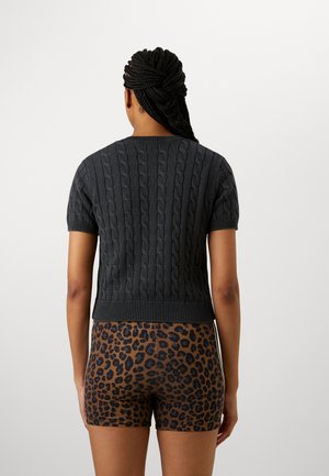 Femme aux cheveux tressés portant un pull court à manches courtes en maille torsadée foncée et un short marron à imprimé léopard, debout de dos à la caméra.