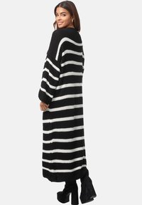 Gestreifter, schwarz-weißer, langer Cardigan mit lockerer Passform, gestrickter Textur, weiten Ärmeln und seitlichen Schlitzen, kombiniert mit schwarzen High-Heels.