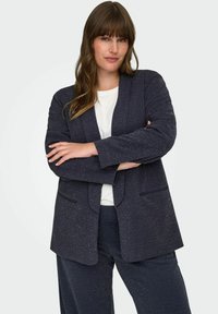 Žena s dlouhými hnědými vlasy v námořnicky modrém sako a kalhotách, bílé košili, stojící se založenýma rukama před jednobarevným pozadím.