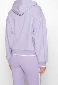 Sudadera recortada en lavanda hecha de una tela suave, con capucha, puños elásticos y un acento de cinturilla blanca. Combinada con leggings a juego.