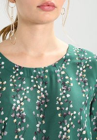 Grünes Blumenoberteil mit einem runden Ausschnitt, versehen mit einem Muster aus weißen, gelben und lila Blumen auf einem glatten Stoff.