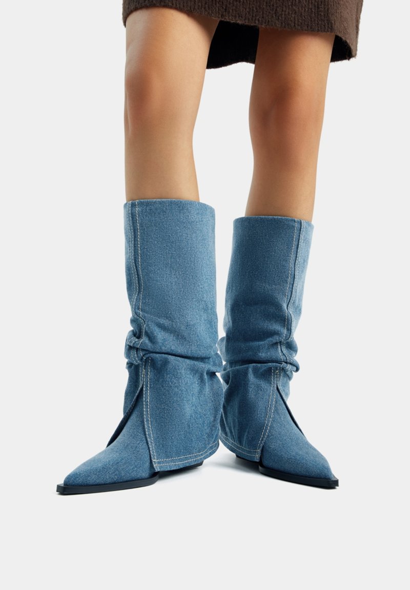 Bottes en denim jusqu'au genou avec un design lâche, bouts pointus et semelles en caoutchouc noire. Tissu bleu clair avec des détails de couture visibles.