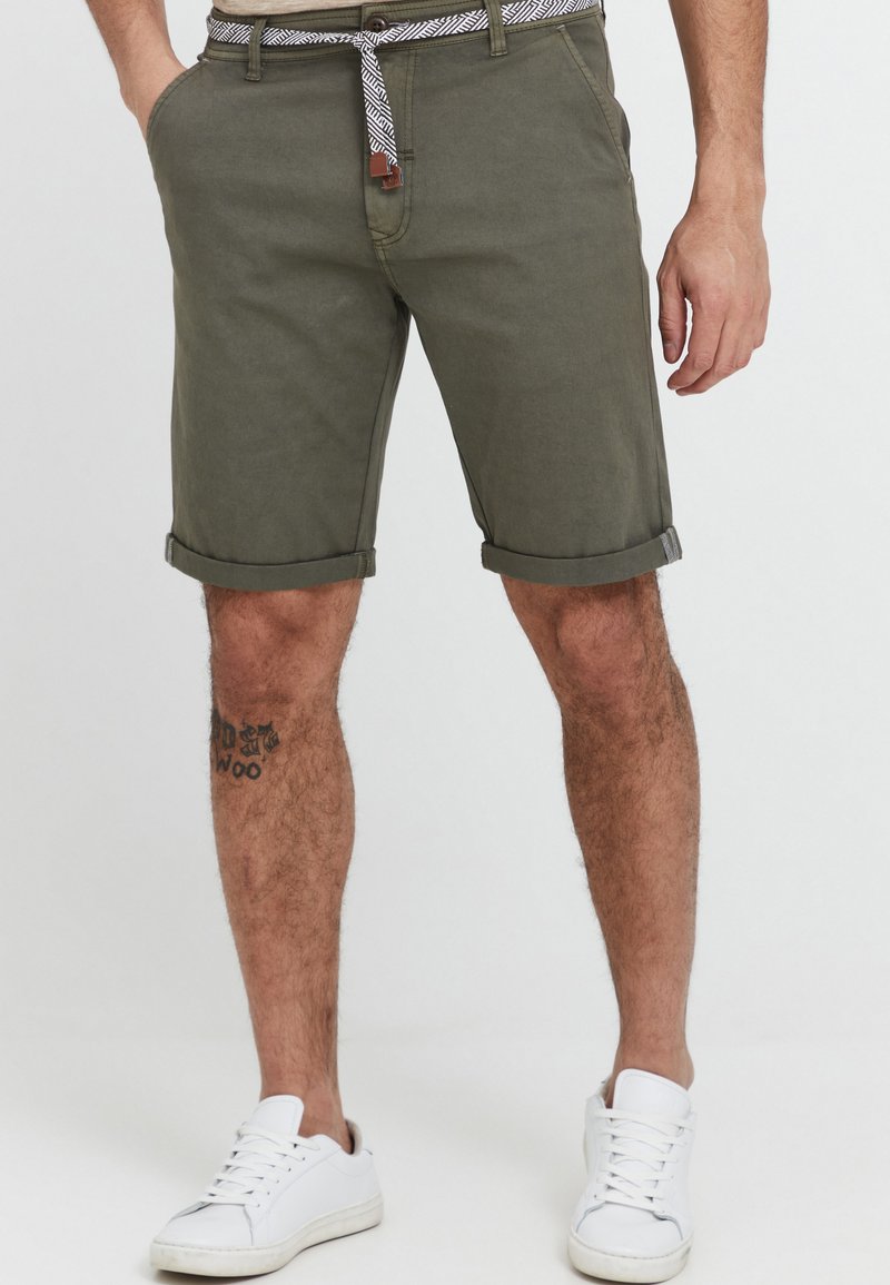 Shorts vert olive à revers, en coton. Présente une ceinture à motifs et des poches latérales. Associés à des baskets blanches.